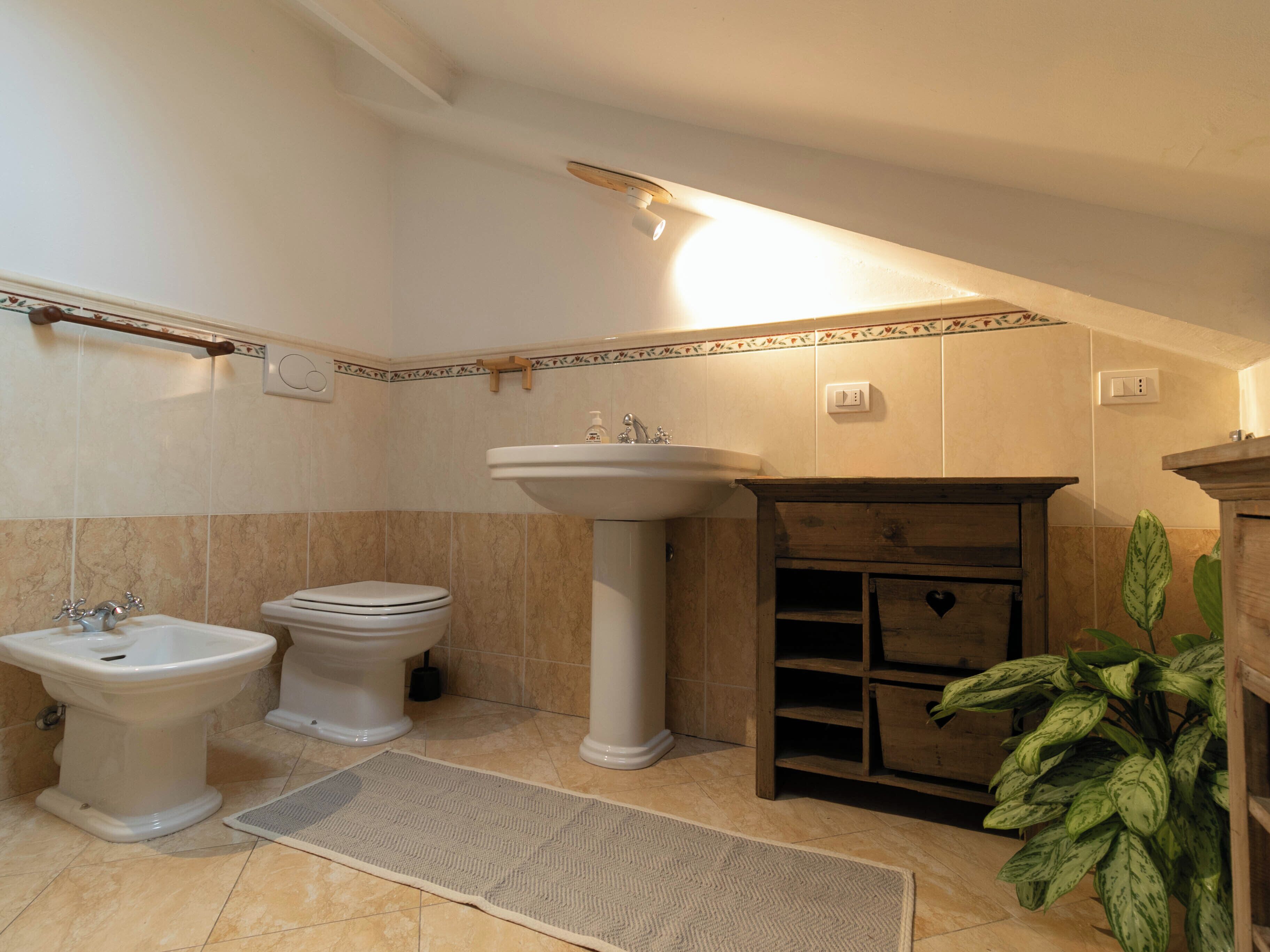 A bathroom with a pedestal sink, a bidet, and a toilet. There is a wooden cabinet with a heart-shaped decoration and a potted plant.
