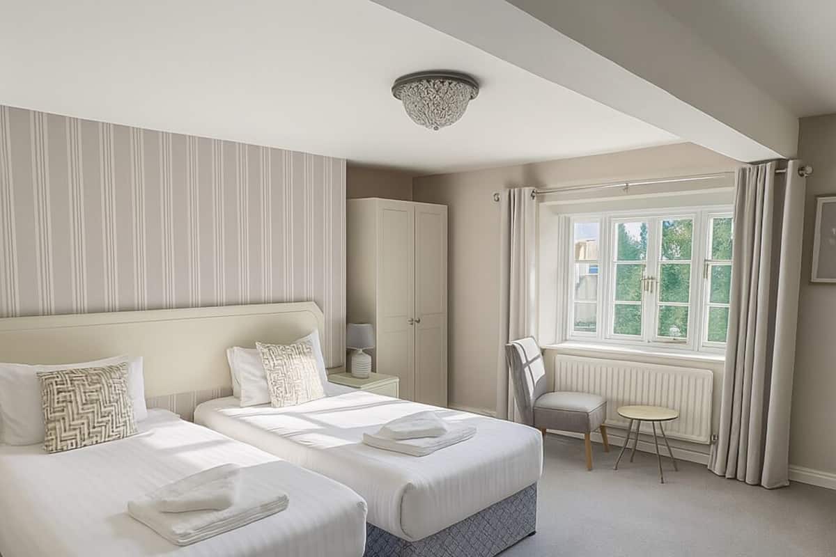 A hotel room with two beds, a desk, a chair, a radiator, and a window with curtains.