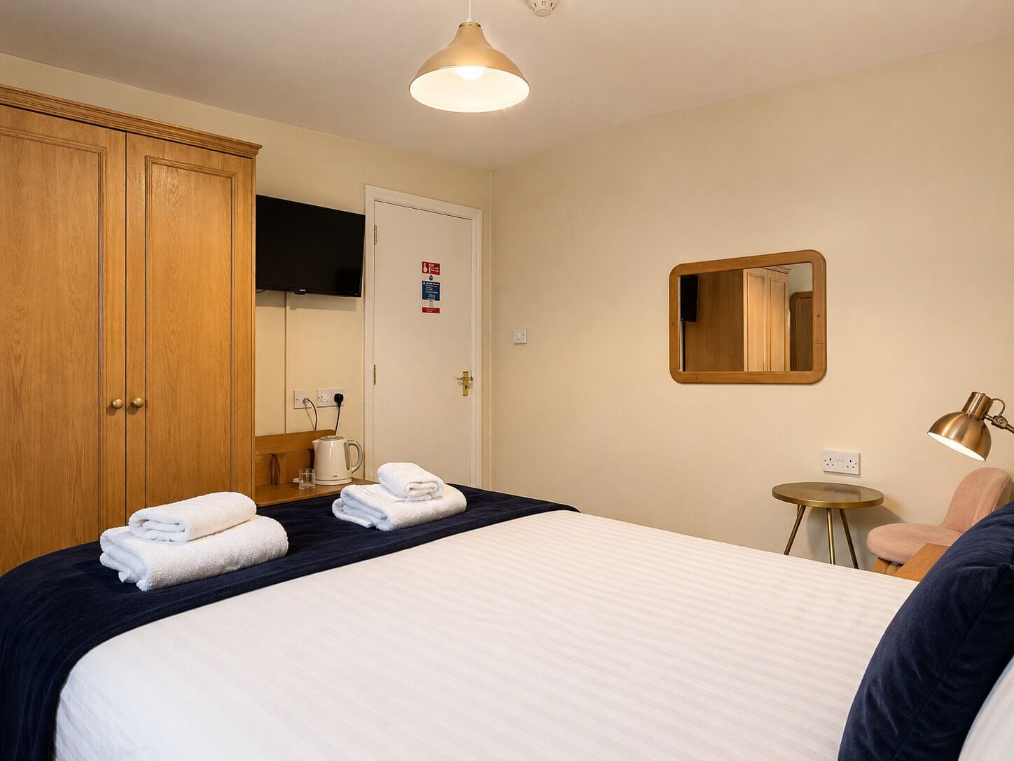 A hotel room with a bed, a wooden wardrobe, a television, a mirror, a small table with a lamp, and a coffee maker.