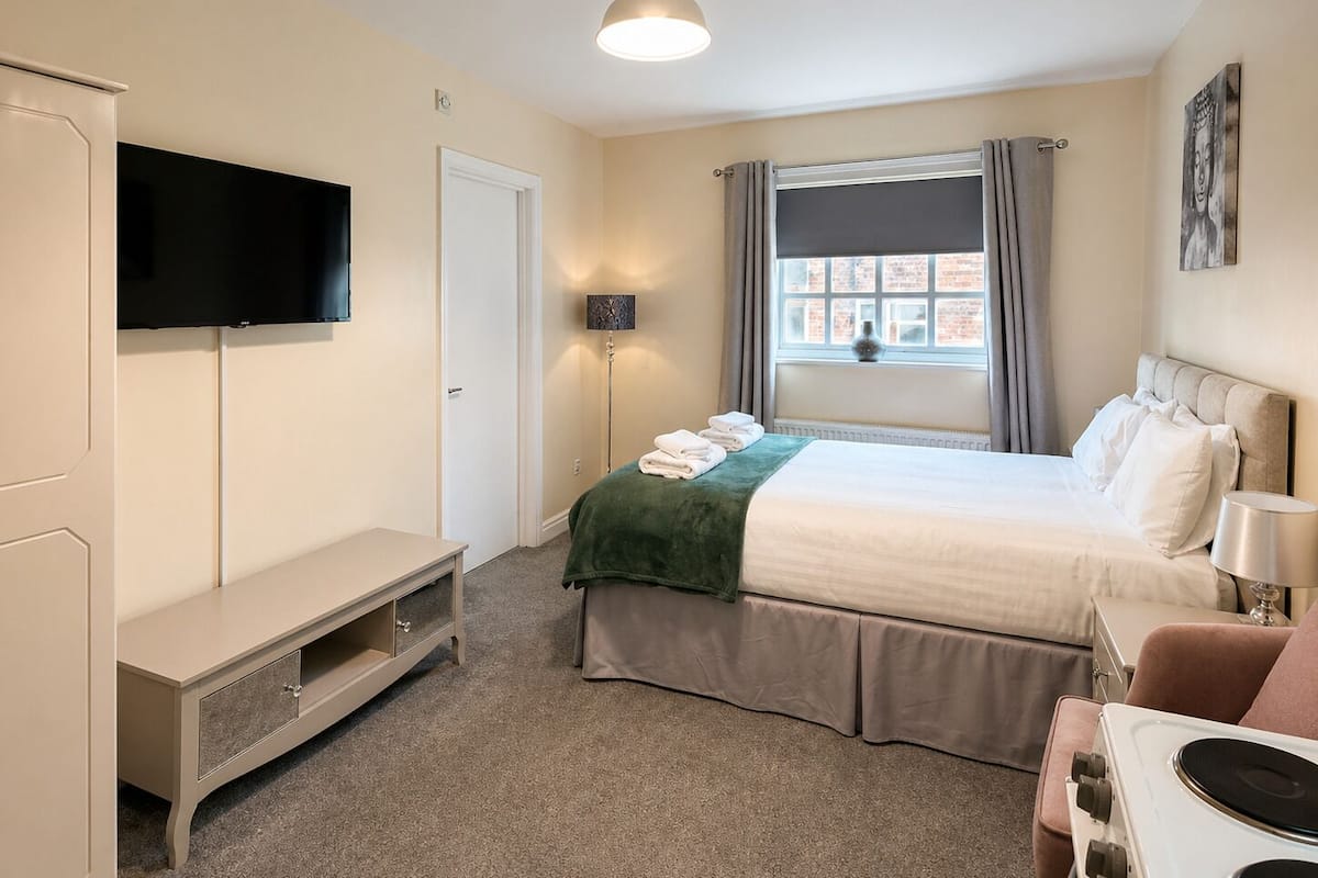 A bedroom with a bed, a TV, a desk, a chair, a lamp, a window with curtains, and a wall-mounted artwork.