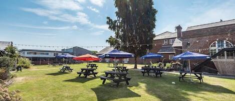An outdoor patio area with picnic tables and umbrellas, surrounded by brick buildings and a large tree.