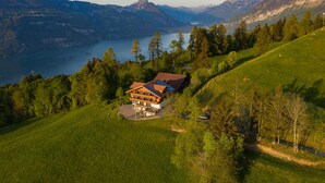 A large, two-story wooden house with a sloped roof, surrounded by lush green fields and trees, overlooking a lake and mountains.
