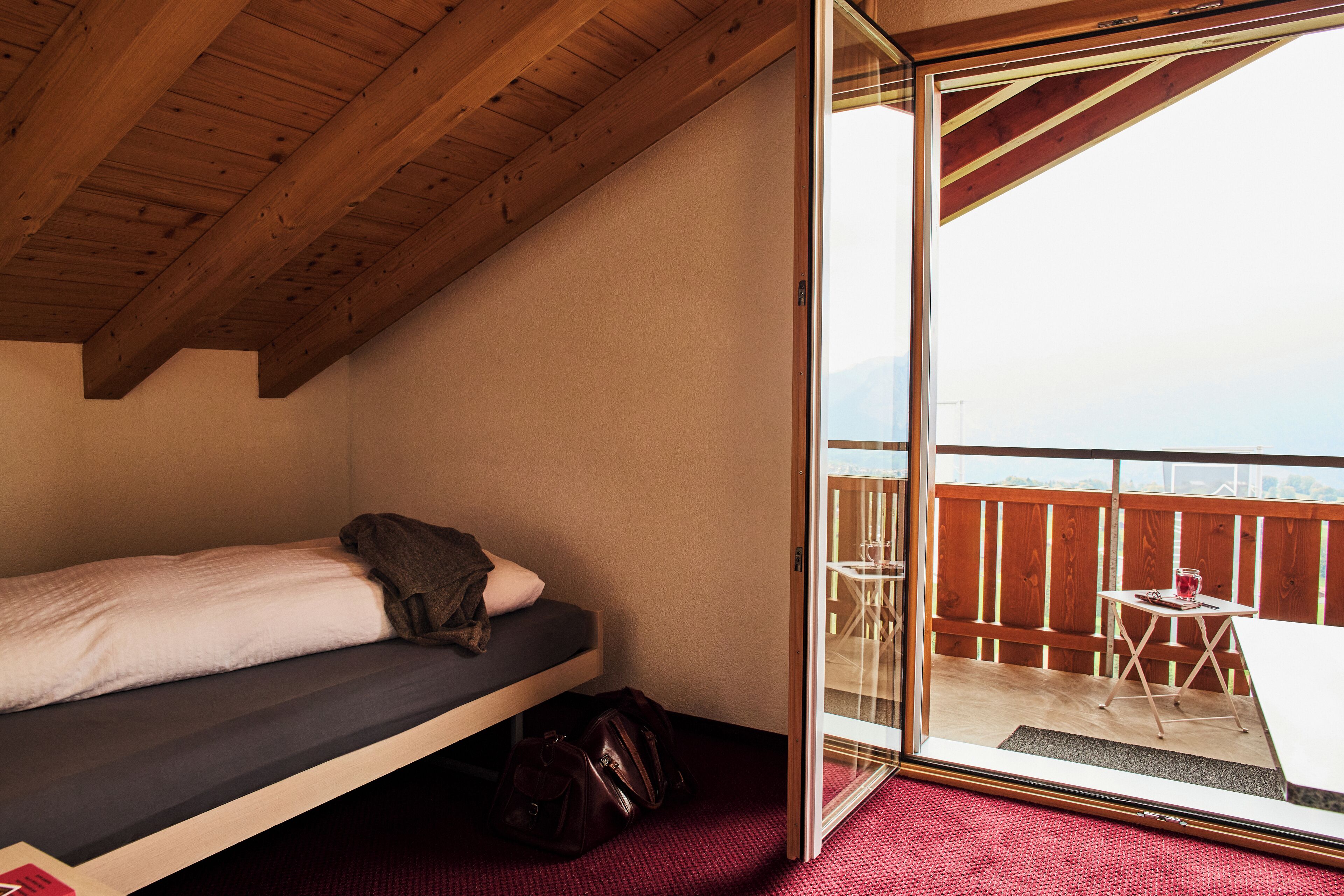 Ein kleines Zimmer mit einem Einzelbett, Holzboden und einer Glasschiebetür, die auf einen Balkon mit Tisch und Stühlen führt.