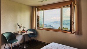 A hotel room with a bed, a table with chairs, and a large window offering a scenic view of mountains and a lake. - Auszeit-Hotel Z Aeschiried (Retreat Hotel Z Aeschiried) (Aeschi bei Spiez)