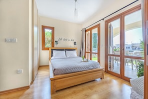 A bedroom with a wooden bed, a view of the outdoors through large windows, and a wall-mounted light fixture. - Aa02 Adjacent to Aphrodite Hills- Communal Pool, Garden (Kouklia)
