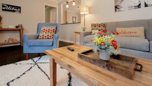 Un salon avec un fauteuil bleu, un canapé gris, une table basse en bois ornée d’un vase de fleurs et un panneau « Bienvenue » accroché au mur.