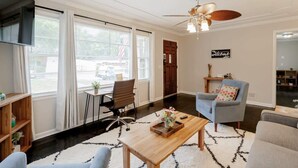 Un salon moderne avec une table basse en bois, un fauteuil bleu et un canapé. On y trouve un ventilateur de plafond, un téléviseur fixé au mur et une fenêtre avec des rideaux.