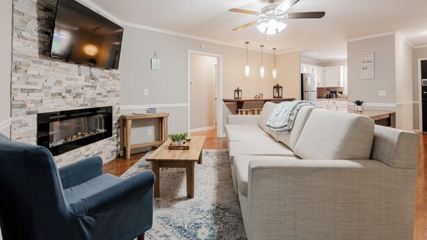 A living room with a fireplace, a TV mounted on the wall, a ceiling fan, and a kitchen in the background.