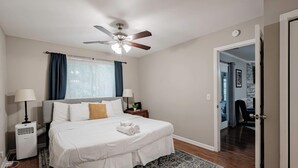 A bedroom with a bed, two bedside tables, a ceiling fan, a window with curtains, a patterned rug, and a door leading to another room.