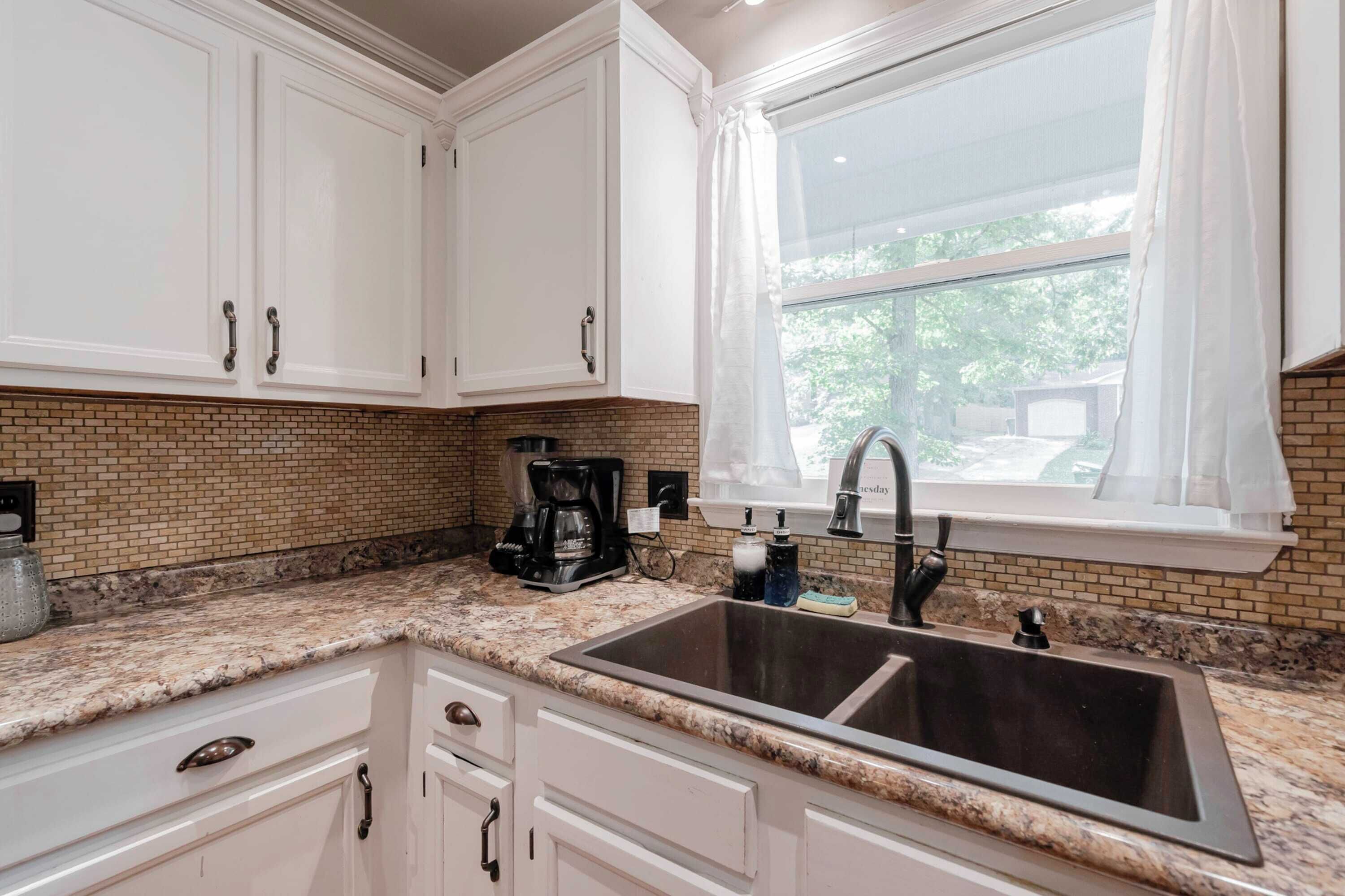 Una cocina con armarios blancos, encimera de granito, pileta doble y ventana con cortinas blancas.