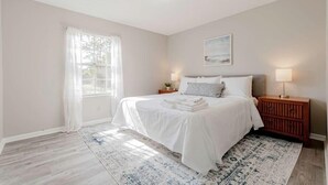 Un dormitorio con cama, mesita de noche, lámpara, ventana con cortinas translúcidas y alfombra estampada.