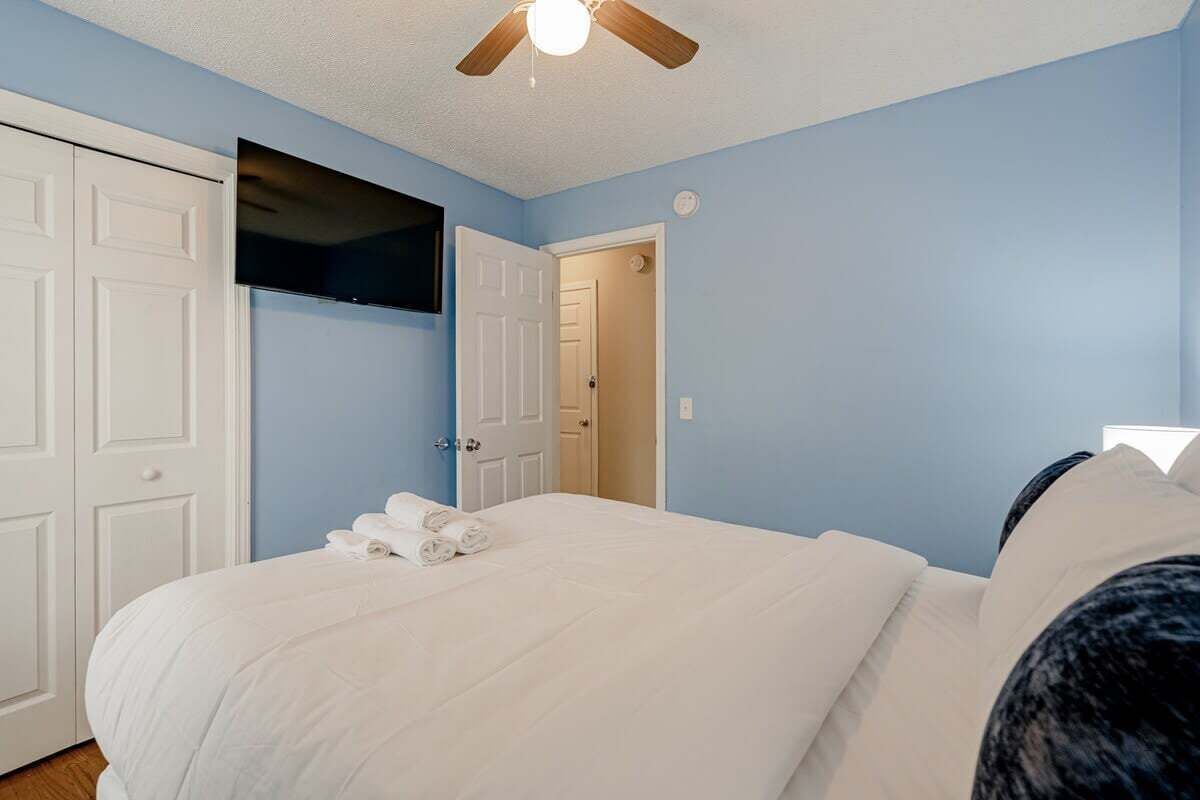 A bedroom with a bed, white walls, a ceiling fan, and a TV mounted on the wall.