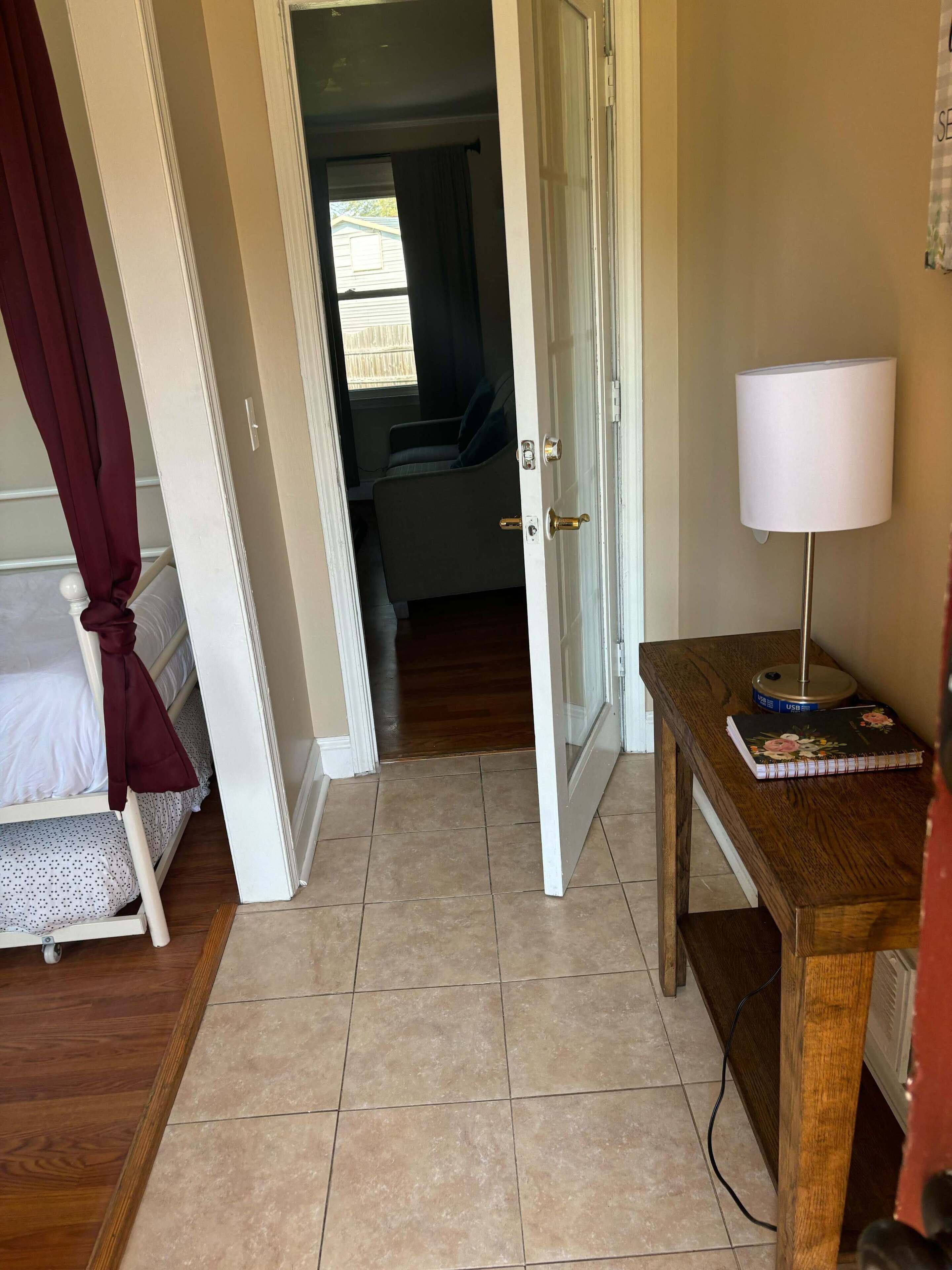 A hotel room with a bed, a desk with a lamp, and a view into the adjacent room.