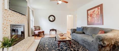 A living room with a grey sofa, a patterned rug, a ceiling fan, a flat-screen TV mounted on a brick wall, and a painting on the wall.