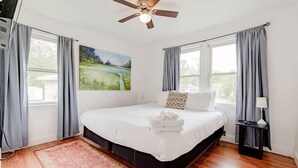 Een slaapkamer met een bed, een plafondventilator, een schilderij, een nachtkastje en een raam met gordijnen.
