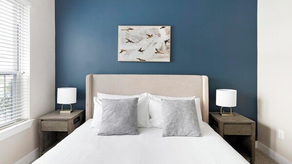 A bedroom with a bed, two bedside tables, a wall-mounted artwork, and a window with blinds.