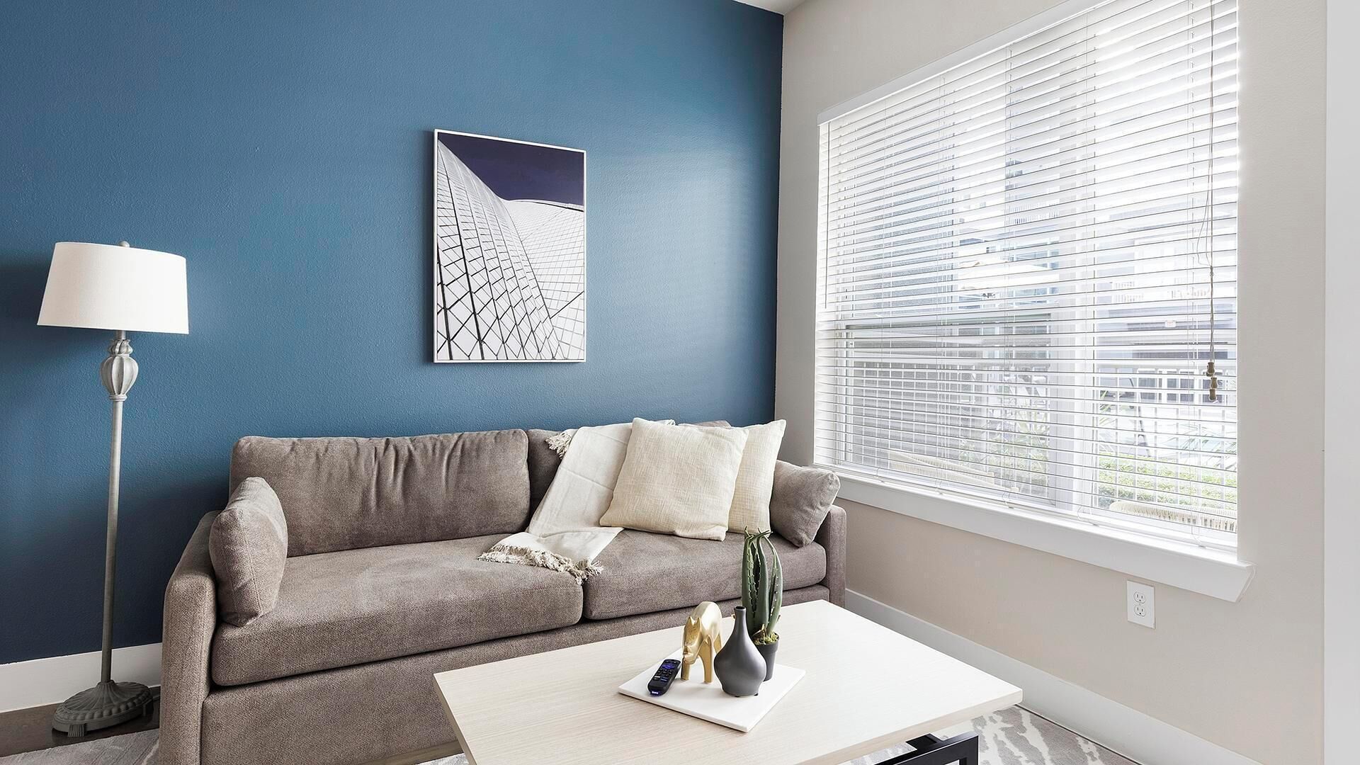 A living room with a grey sofa, a white coffee table, a floor lamp, and a window with blinds.