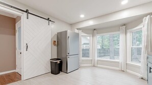 A modern kitchen with a sliding door, a refrigerator, a trash bin, and large windows offering a view of the outdoors. - Mins to Lake Murray & Downtown - The Settle-Inn (Irmo)
