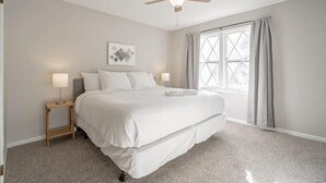 Une chambre à coucher avec un lit, un ventilateur de plafond, une fenêtre avec des rideaux et une petite table de chevet avec une lampe.