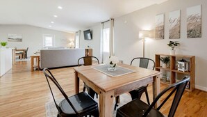 Uma sala de estar moderna com uma mesa de jantar de madeira, cadeiras pretas, um sofá cinza e uma área de cozinha ao fundo.