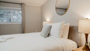 Uma cama bem arrumada com lençóis brancos e travesseiro cinza, uma mesa de cabeceira com abajur e um espelho redondo na parede.