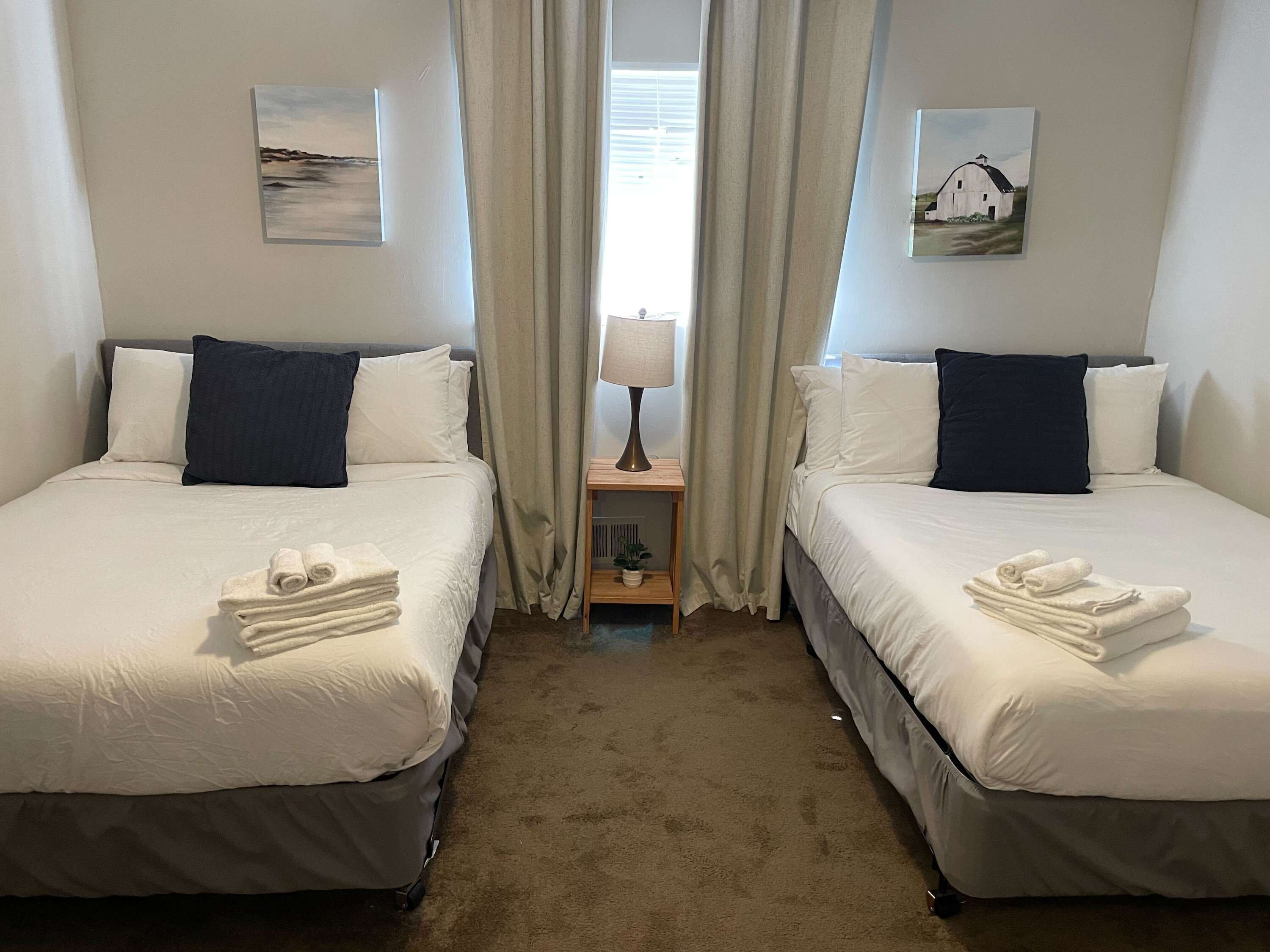 Ein Hotelzimmer mit zwei Betten, jeweils mit weißer Bettwäsche und dunklen Kissen. Auf den Betten liegen Handtücher, ein kleiner Tisch mit einer Lampe und Vorhänge.