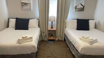 Ein Hotelzimmer mit zwei Betten, jeweils mit weißer Bettwäsche und dunklen Kissen. Auf den Betten liegen Handtücher, ein kleiner Tisch mit einer Lampe und Vorhänge.