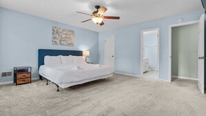 Quarto com cama, mesa de cabeceira, ventilador de teto e porta para o banheiro.
