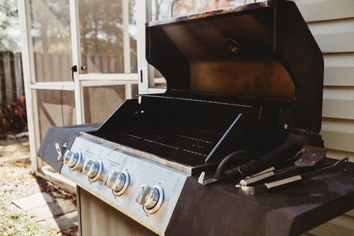 Una parrilla para barbacoa con cuatro perillas de control y una rejilla de cocción negra, frente a una casa con ventana y una cerca de madera al fondo.