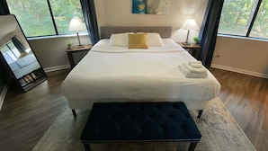 Quarto de hotel com uma cama grande, duas mesas de cabeceira com luminárias, um quadro na parede e grandes janelas com cortinas.