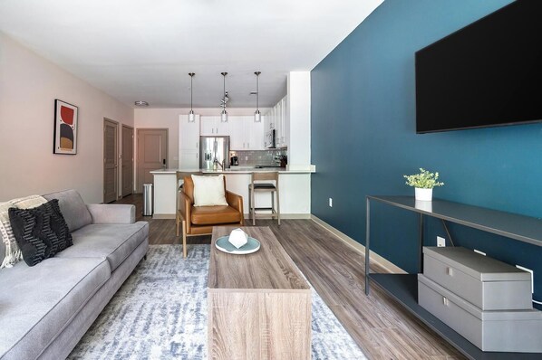 Una sala moderna con un sofá, una mesa de centro y una silla. Hay una zona de cocina con armarios y un refrigerador. Un televisor está montado en la pared, y hay una planta en un estante.