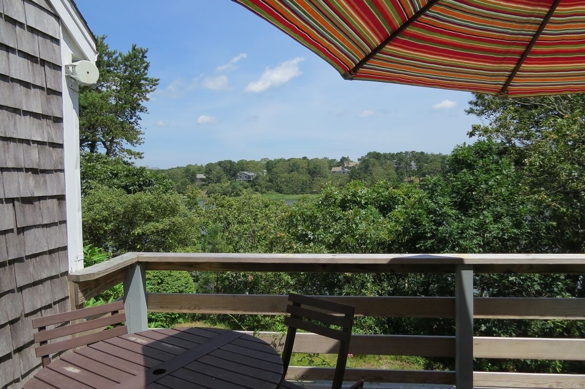 A wooden deck with a table and chair, overlooking a lush green landscape with trees and houses in the distance.