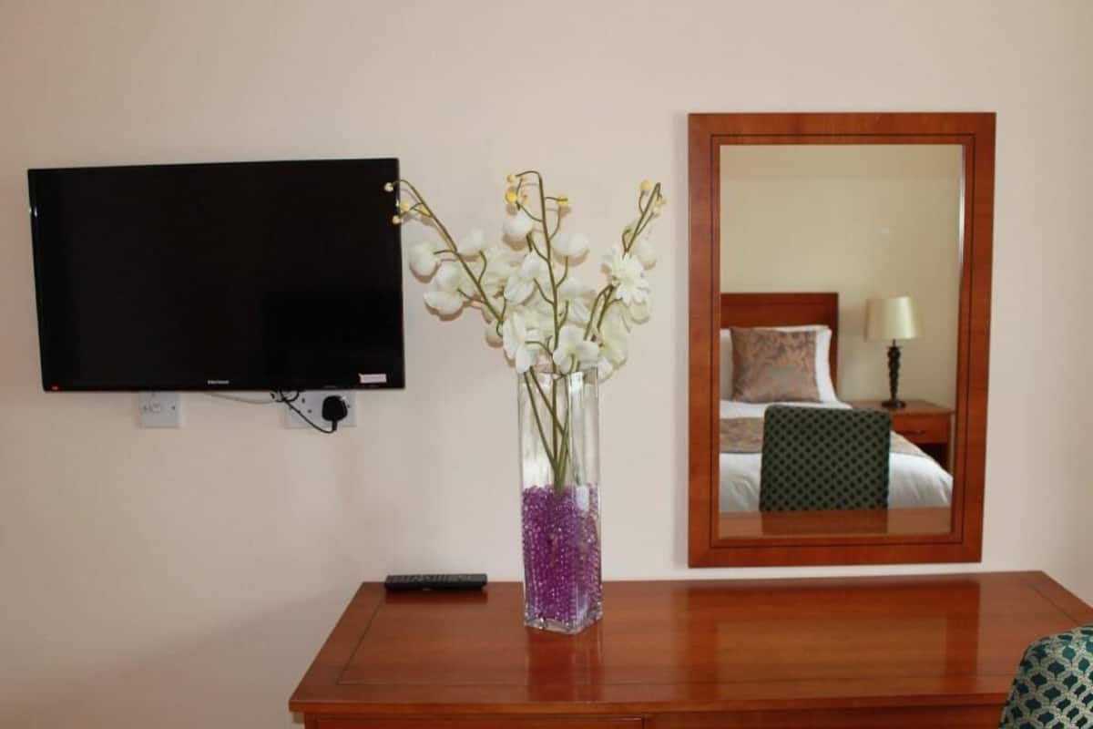 A hotel room with a flat-screen TV mounted on the wall, a wooden bedside table with a vase of white flowers, and a mirror reflecting the room's bed and nightstand.