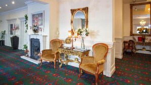 Una habitación con alfombra estampada, una chimenea, dos sillas ornamentales, una mesa con un florero y un espejo grande.