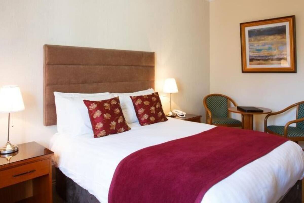 A hotel room with a large bed, two bedside lamps, a nightstand, a desk with two chairs, and a framed picture on the wall.