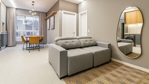 A modern living room with a grey sofa, a dining table with chairs, a large window, and a mirror on the wall. - Gastronomy | Hot Tub | Gym #TRINA04 (Florianópolis)