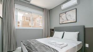 A neatly made bed with a grey headboard, a window with curtains, and a wall-mounted air conditioner. - Gastronomy | Hot Tub | Gym #TRINA04 (Florianópolis)
