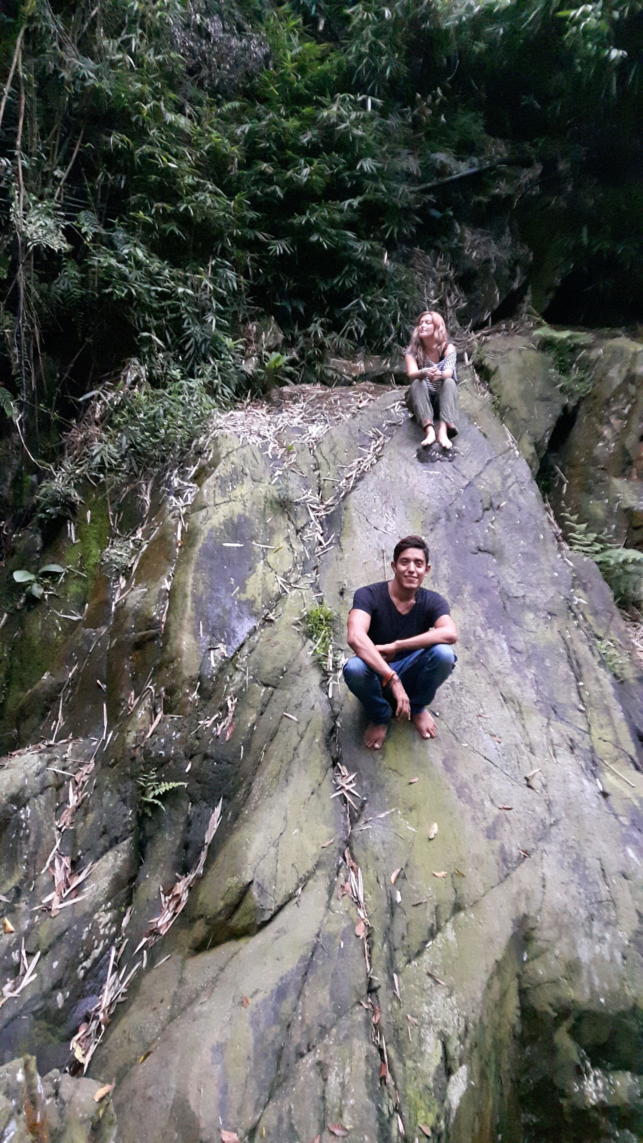 Two people sitting on a rocky surface in a natural setting with dense foliage in the background.