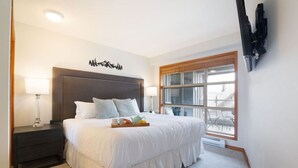 A hotel room with a large bed, a nightstand, a window with a view, and a mounted television. - Ski in/out Aspens 2BR - 3 HotTubs, Pool, Ski Valet, AC (Whistler)