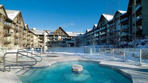 A pool area with a hot tub, surrounded by buildings with balconies and snow on the ground. - Ski in/out Aspens 2BR - 3 HotTubs, Pool, Ski Valet, AC (Whistler)
