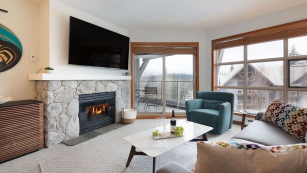 A modern living room with a stone fireplace, a flat-screen TV, a sofa, and large windows offering a view of a snowy landscape. - Ski in/out Aspens 2BR - 3 HotTubs, Pool, Ski Valet, AC (Whistler)
