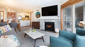A modern living room with a fireplace, a flat-screen TV, and a dining area with a kitchen in the background. - Ski in/out Aspens 2BR - 3 HotTubs, Pool, Ski Valet, AC (Whistler)