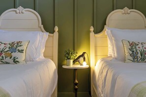 Une chambre d’hôtel avec deux lits, chacun doté d’une tête de lit blanche et de coussins à motifs floraux. Une petite table avec une lampe et une plante en pot se trouve entre les deux lits.