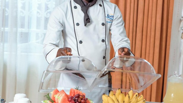 A chef in a white uniform with a chef's hat, serving fruit and bananas from a buffet table. - Jevine Hotel (Kampala)