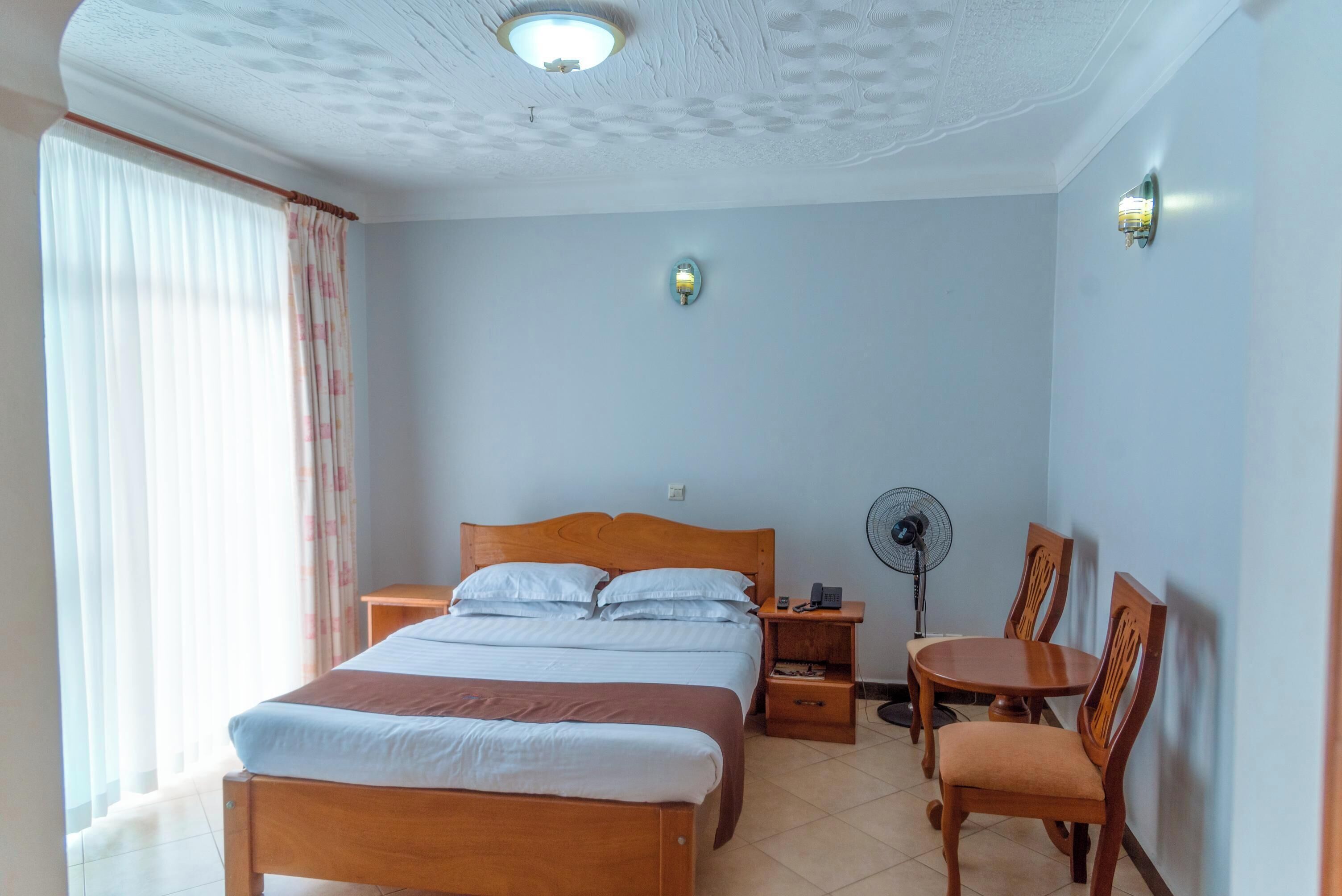 Habitación de hotel con cama de madera, una mesita de noche, una silla, un ventilador de pie y una pequeña mesa.