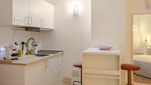 A compact kitchen with white cabinets, a sink, and a countertop with a coffee machine. There is a small dining area with a table and two stools.