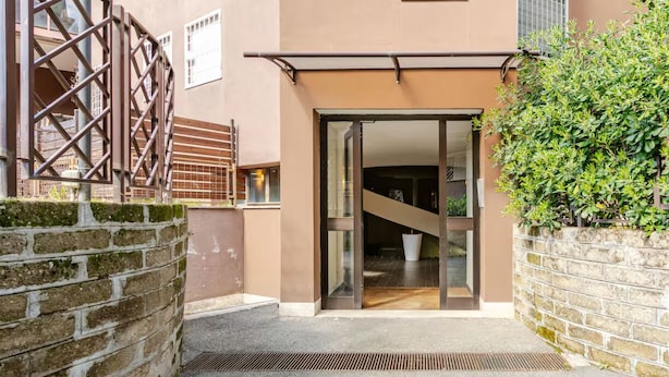 A building entrance with a glass door, a staircase leading inside, and a brick wall with greenery.