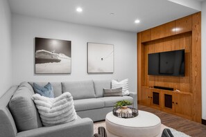 A modern living room with a grey sofa, a round coffee table with a plant, and a flat-screen TV mounted on a wooden wall.
