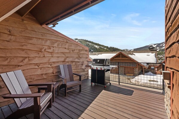 A wooden deck with Adirondack chairs and a grill, overlooking a snowy landscape and buildings.