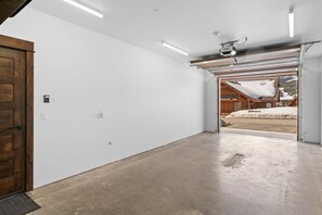 A spacious garage with a concrete floor, a wooden door, and a view of a snowy landscape through an open garage door.